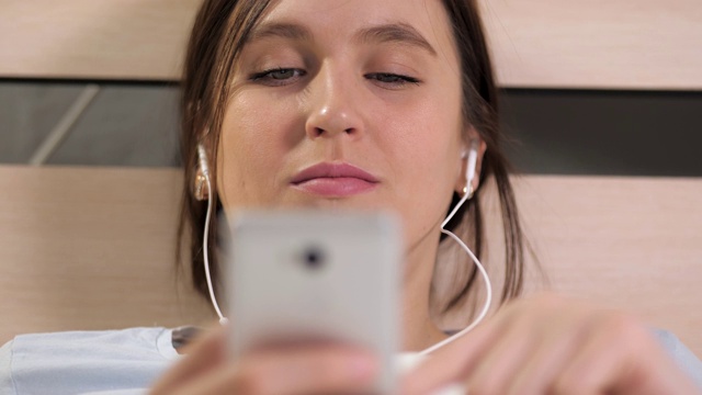 女孩听音乐和使用电话。年轻迷人的女子躺在床上，戴着耳机听着音乐，一边翻阅手机屏幕。特写镜头视频素材