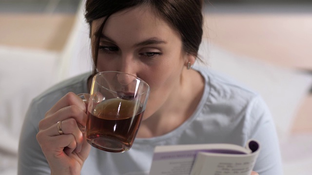 女孩正在读书。年轻貌美的白人女子晚上坐在床上看书，睡前喝红茶。特写镜头视频素材