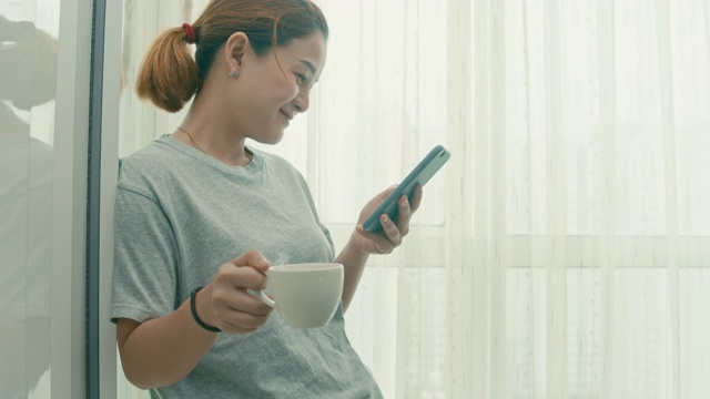 一名女子站在窗边，一边用智能手机一边在家喝热咖啡视频素材