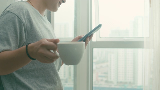 女人站在窗户附近，用智能手机喝热咖啡，特写视频素材