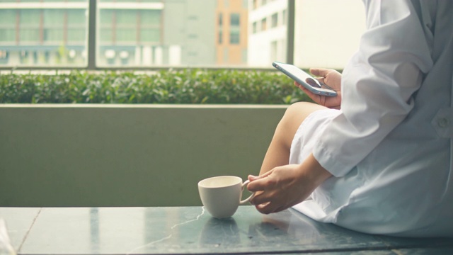 女医生站在窗口边用手机边喝热咖啡视频素材