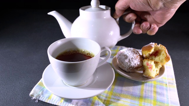 Black tea pouring from teapot into cup. Sweet cakes are on a table.视频素材