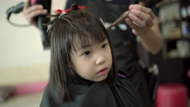 美发师在美容院用吹风机吹干小女孩的头发。发廊里的亚洲小女孩。小女孩拥有幸福，享受头发护理。手持拍摄，真实生活视频下载