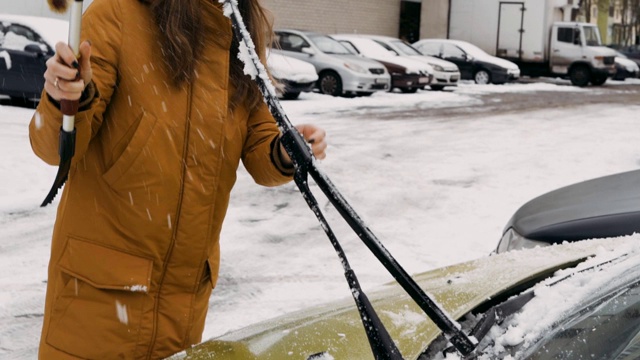 在院子里，一个穿着温暖棕色夹克的女人在清理雨刷上的雪视频素材