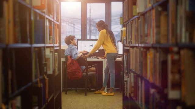 大学图书馆:男孩在书桌上使用个人电脑，与女同学交谈，女同学解释，帮助他完成课堂作业。专注的学生一起学习。在一排排书架之间拍摄视频素材