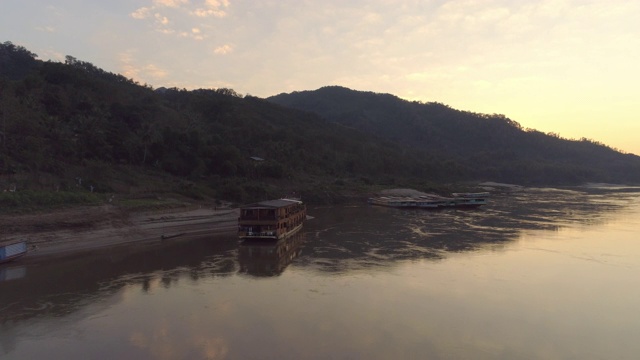 航拍邮轮在河岸附近的树木在森林对天空，无人机飞向前越过河流向山脉日落-湄公河，老挝视频素材