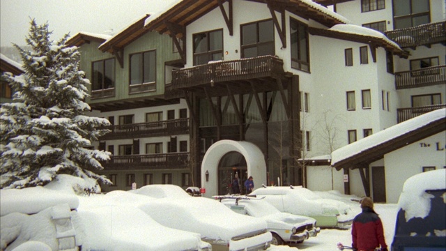 多层白色与棕色修剪滑雪小屋在下雪的日子。看起来像维尔滑雪胜地。在前景停车场看到被雪覆盖的汽车。见左侧的针叶树被雪覆盖。看到行人在下雪时走过。放大到入口上方的阳台。视频素材