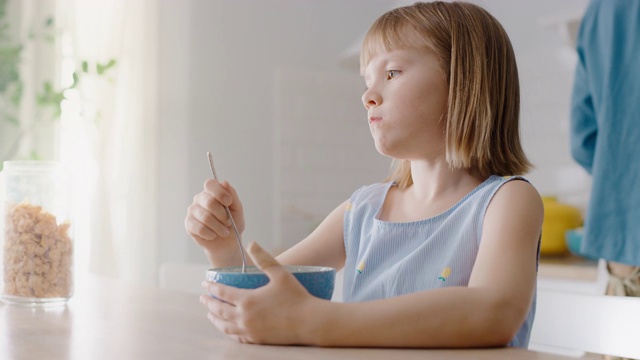 厨房里的早餐:可爱的小女孩吃着健康的格兰诺拉麦片，碗里盛着牛奶。她可爱的小女孩吃早餐。背景:关怀备至的母亲在准备食物视频素材