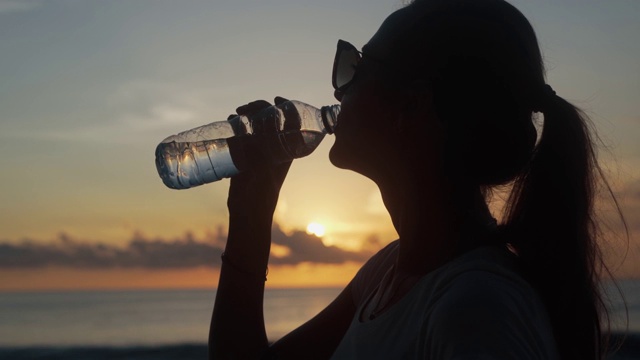 日落时分，一个戴着墨镜的女人从瓶子里喝清水视频素材