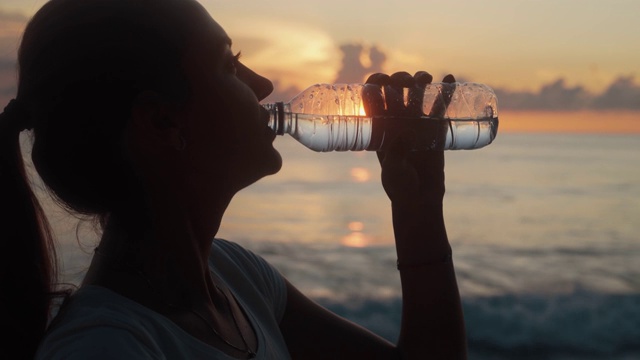 一个女人的剪影喝干净的水从瓶子在日落在海洋海滩视频素材