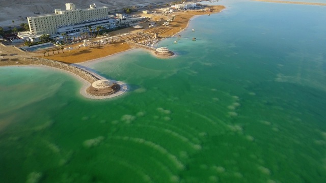 在阳光明媚的一天，无人机在著名的绿松石盐湖上空向前飞行，航拍死海海滩上的酒店视频素材