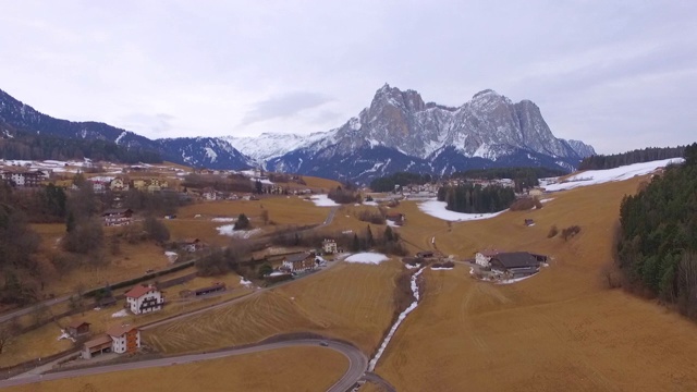 空中拍摄的雪在岩石山附近的村庄与天空，无人机从住宅建筑向后飞行在冬季- Dolomites，意大利视频素材