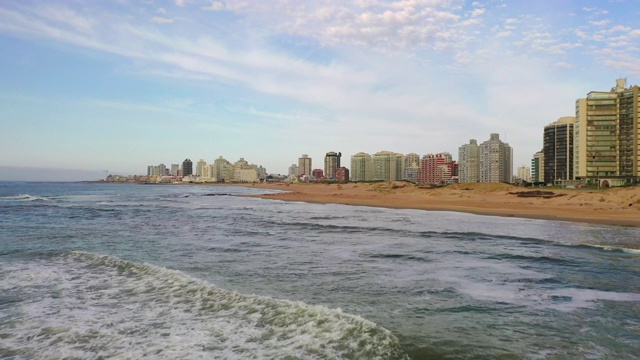 航拍城市建筑附近的海浪溅起浪花，无人机迎着天空飞向海滩——乌拉圭埃斯特角视频素材