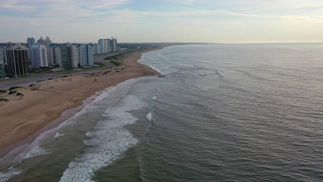 航拍海浪在海滩上溅起浪花的城市，无人机向前飞行的海岸线对天空-埃斯特角，乌拉圭视频素材