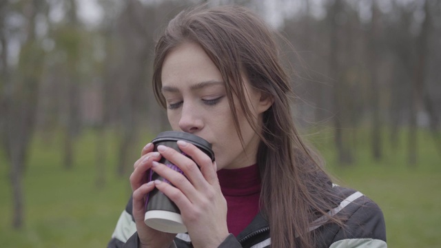 体贴迷人的女孩在户外喝咖啡的特写。美丽的黑发女人与棕色眼睛享受阴天在公园的肖像。生活方式，休闲，休息，自然视频素材