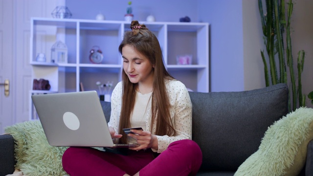 一个女人连接着一台笔记本电脑，用信用卡在网上购物。年轻女孩坐在沙发上用信用卡在网上购物。女性使用笔记本电脑进行在线交易视频素材