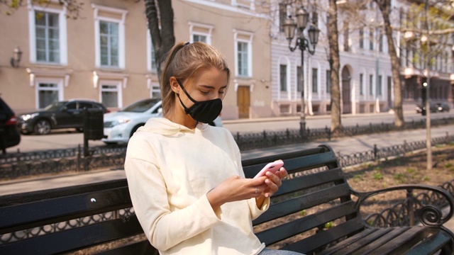 年轻迷人的女人戴着防护面具坐在长椅上使用智能手机在老城区中心的街道上，慢镜头视频素材