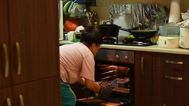 一位亚洲华人中年妇女从厨房的烤箱中取出蛋糕托盘，准备做饭视频素材