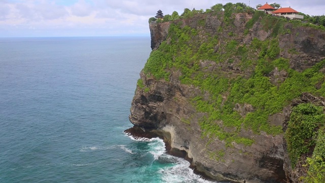 空中静态的黄昏海面，平静的波浪翻滚在岩石海岸。惊人的高悬崖之上的海洋和顶部的乌鲁瓦图寺庙。印尼巴厘岛视频素材