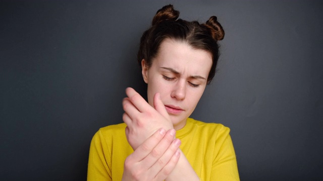 近距离的不幸的年轻女子痛苦的手或手腕，穿着黄色毛衣，孤立在灰色的背景。腕管综合征，关节炎，神经系统疾病的概念。视频素材