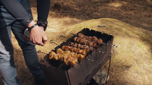 男用手在户外烧烤架上的烤肉上喷啤酒。野餐时用木炭烧烤肉。视频下载