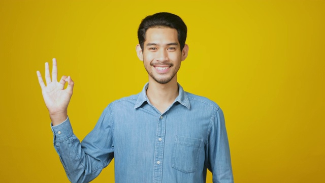 年轻的亚洲男子微笑着显示他的ok手势，快乐的亚洲男子站在工作室的黄色背景上，看着相机做出ok手势，头部拍摄，4k分辨率视频素材