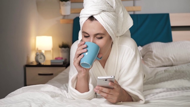 女孩喝咖啡或茶和使用电话。漂亮的女人在卧室里穿着白色的浴袍躺在床上喝着热咖啡或茶，在手机触摸屏的屏幕键盘上输入文字。特写镜头视频素材