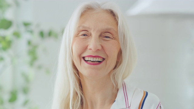 居家一间明亮的房间里的老年女性老人的真实美丽画像镜头。美丽的老女人与灰色头发摆好姿势的相机和温柔的微笑。健康快乐的老人。视频素材