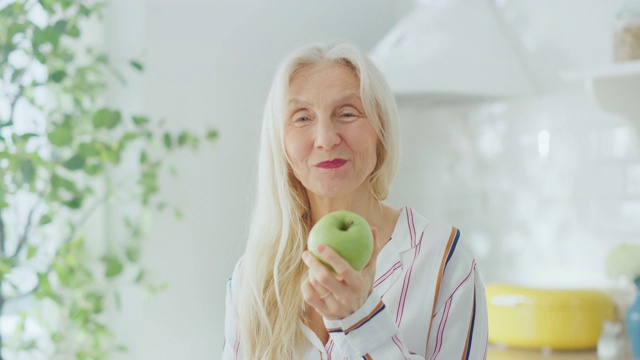 真实的女退休老人在一个明亮的厨房里吃苹果。美丽的老女人与灰色头发摆好姿势的相机和温柔的微笑。快乐素食充满健康。视频素材