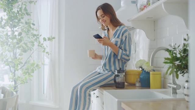 美丽的女性坐在橱柜上，在厨房里使用智能手机，同时喝着一杯新煮的咖啡。穿着睡衣，有着健康生活方式的女孩早上在家里放松。视频素材