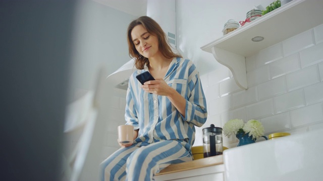 美丽的女性坐在橱柜上，在厨房里使用智能手机，同时喝着一杯新煮的咖啡。穿着睡衣的年轻女子收到朋友的短信后笑了。视频素材