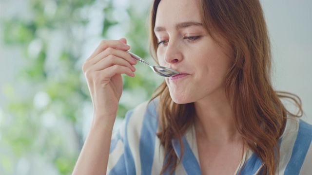 一个美丽的年轻女性在家里用勺子吃奶油酸奶的真实肖像。迷人的黑发女人为相机摆姿势和温柔的微笑。拥有健康生活方式的快乐的人。视频素材