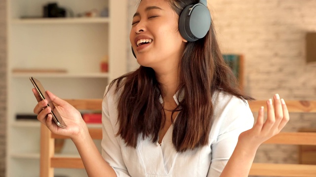 快乐的年轻亚洲女人戴着耳机唱歌和听音乐，而坐在床上，在家里拿着手机视频素材