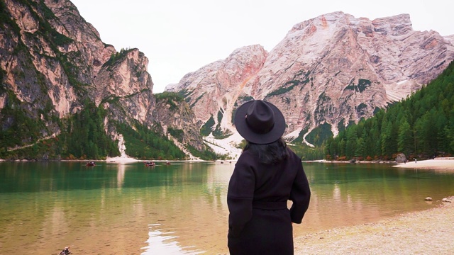 布雷斯湖畔的一个女人。梦想成真。享受看旅游风景如画的意大利北部白云石阿尔卑斯山脉。时尚的黑色保暖外套帽子宽边黑色头发时尚模特。转身背向视图视频素材