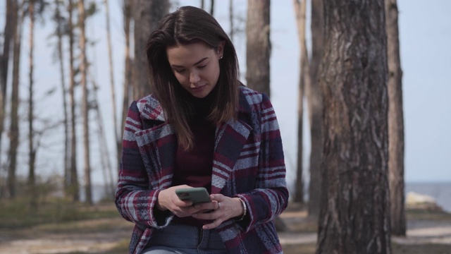 集中的白人女孩在森林里使用智能手机的肖像。黑发的千禧女性在户外上网。无线技术，休闲，生活方式，自然。视频素材