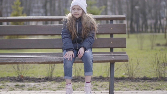 快乐的小女孩坐在长椅上聊天。阳光明媚的日子里，一个可爱开朗的白人小孩在公园里做鬼脸。快乐，乐趣，休闲，生活方式视频素材