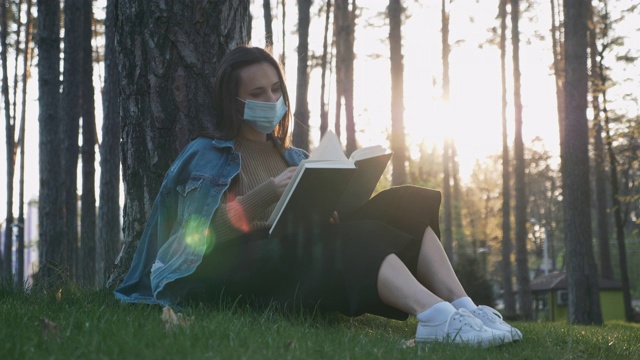 女孩戴着医用防护口罩，在美丽的夕阳下坐在公园的草地上放松和看书。在隔离期间，可爱的女人戴着医用口罩在公园看书视频素材