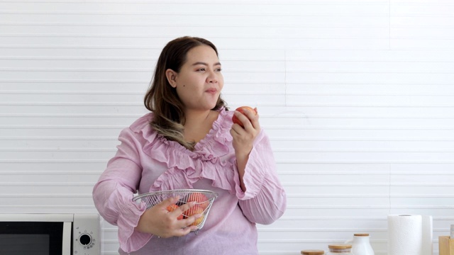 快乐的胖女人站在厨房里吃着美味的苹果。胖女人有健康水果和减肥食品以及低热量水果的概念视频素材