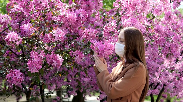 一个戴着医用口罩的女孩站在一棵开花的树旁。冠状病毒流行。新型冠状病毒肺炎视频素材