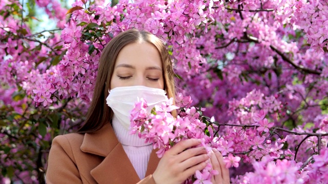 一个戴着医用口罩的女孩站在一棵开花的树旁。冠状病毒流行。新型冠状病毒肺炎视频素材