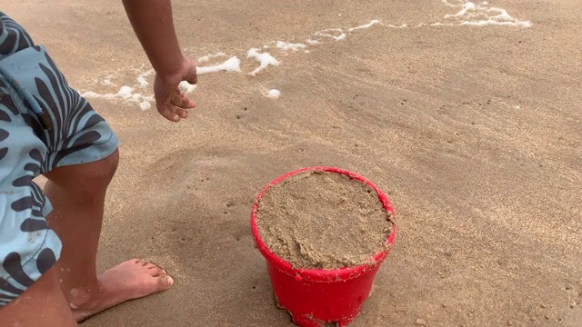 一个小男孩背着装满沙子的桶在海滩上建造沙堡视频素材