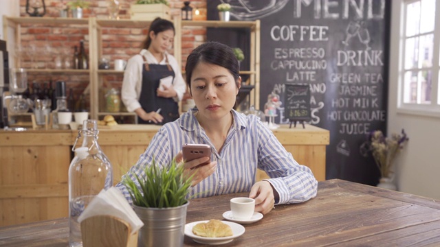 在柜台工作的女咖啡师。亚洲女性顾客在手机上浏览网页。一个女孩在咖啡店里拿着热咖啡，皱着眉头，感觉很奇怪，很难闻视频素材