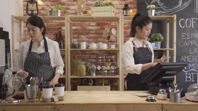 女店员戴着围裙在餐厅为顾客点餐后用平板电脑。两个女孩同事走进柜台准备咖啡。小型创业公司的合伙人。视频素材