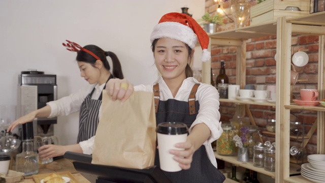 微笑的年轻女咖啡师在围裙拿着咖啡去杯和带走食物在纸袋面对相机。两个咖啡馆酒吧的女店员在圣诞假期戴着圣诞帽在店里工作。视频素材