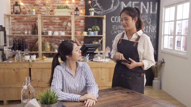 年轻快乐的女服务员穿着牛仔围裙，用平板电脑在咖啡厅里为女顾客点菜。一位办公室女士在等咖啡的时候用手机。友好的女咖啡师工作。视频素材