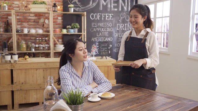 年轻的办公室女顾客坐在桌边与穿着围裙的女服务员聊天。咖啡师端着托盘盘子后给朋友送牛角面包和咖啡。咖啡厅工作人员在酒吧与客户聊天视频素材