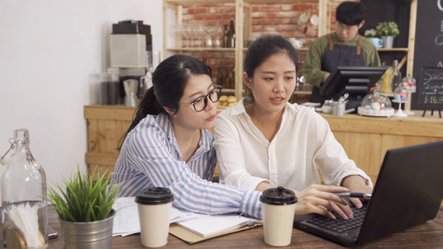 两个年轻的女工在一家餐馆的咖啡厅里碰面。美丽的办公室女同事在咖啡馆酒吧的笔记本电脑工作时大笑。男服务员站在柜台后面视频素材