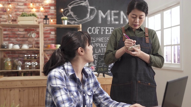 微笑的亚洲女性自由职业者打字在笔记本电脑上订购咖啡从女服务员在咖啡馆。穿着围裙的女士在酒吧写客户订单。餐桌服务对话店店概念视频素材