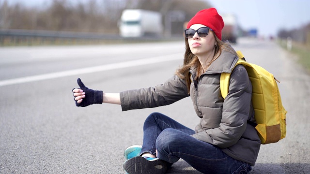 年轻漂亮的女人搭便车，竖起手指。坐在路边视频素材