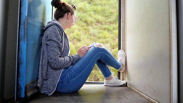 年轻女子坐在打开的火车门附近，看着外面美丽的自然。女性游客乘坐客运铁路，享受夏日旅游。假期的概念。视频素材